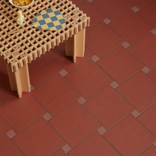 Terracotta red square floor tiles with dark grout lines and small raised decorative squares at intersections, shown in a styled interior scene with a wooden table and tabletop accessories.