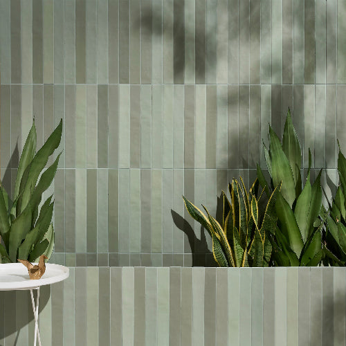 Vertical rectangular wall tiles in a muted sage green and gray colorway, styled with potted green plants and a small white side table in the foreground.