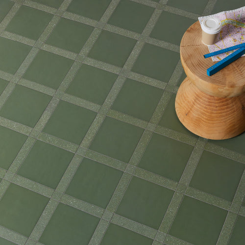 A tile with a rough texture featuring a geometric pattern in various shades of green, placed on a wooden stool with a map and a compass in the background.
